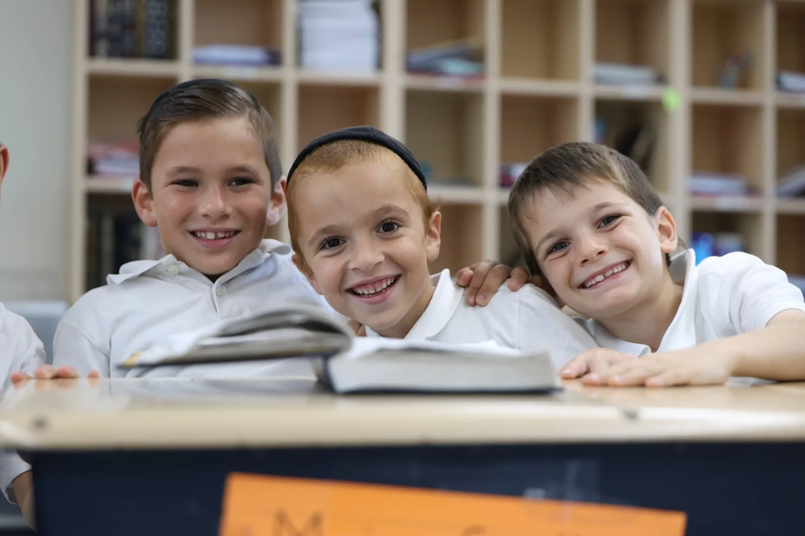 Three boys smiling together at a desk with an open sefer